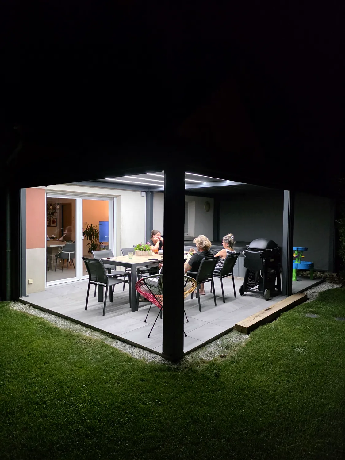 Pergola bioclimatique anthracite de nuit, lames fermées et LED allumées au-dessus d’une table dressée sur une terrasse dallée.