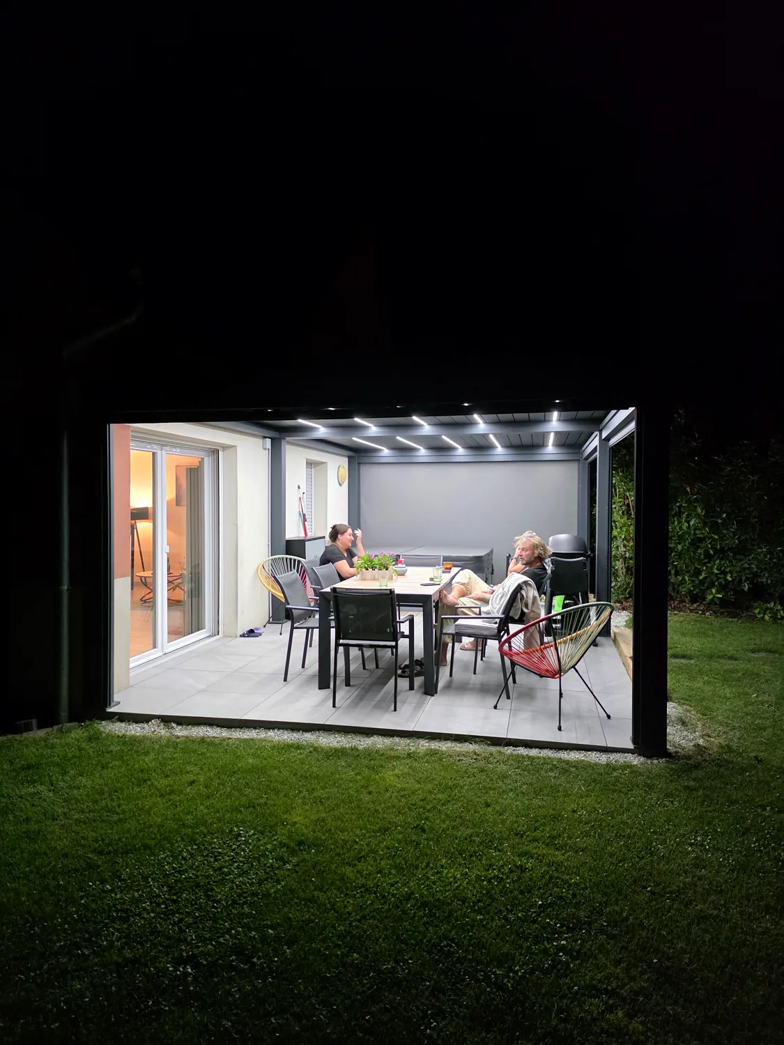 Pergola bioclimatique anthracite de nuit, store latéral baissé, table dressée sous les LED sur une terrasse dallée.