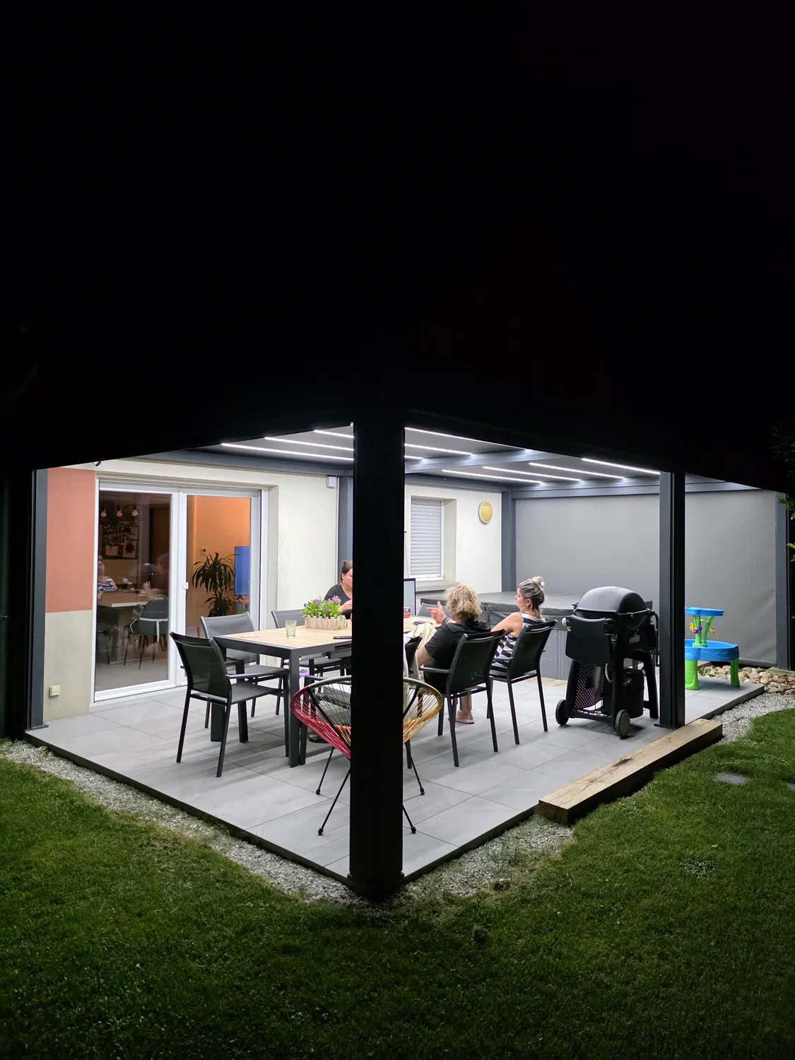 Invités attablés sous une pergola bioclimatique éclairée par les LED intégrées aux lames, terrasse dallée, baie vitrée ouverte sur la maison.