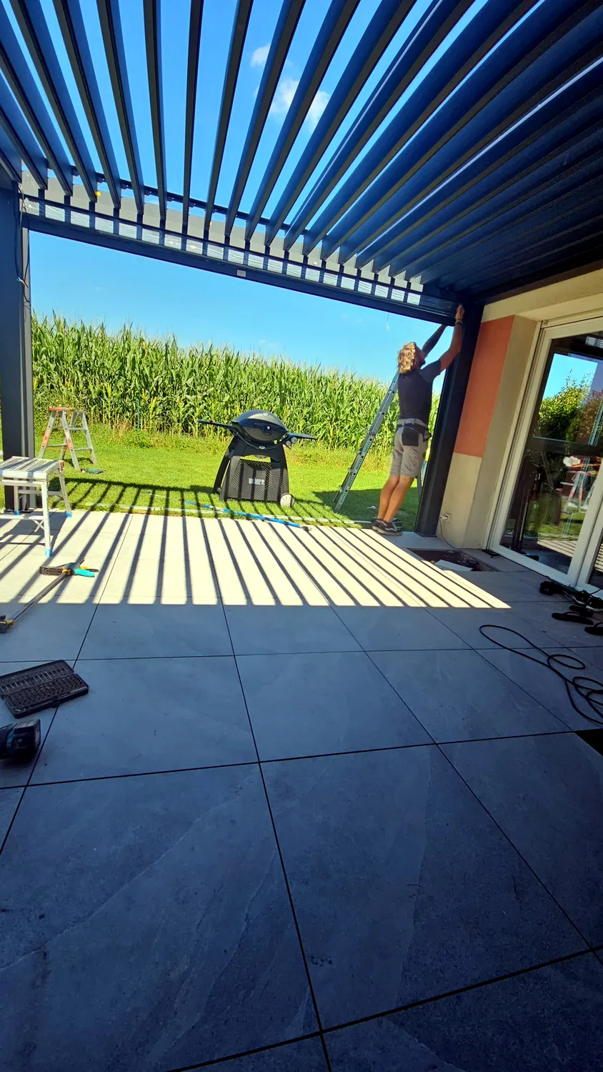Lames bioclimatiques ouvertes en plein soleil, ombres parallèles sur la terrasse carrelée, poseur sur un escabeau contre la façade, champ et ciel dégagé au fond.