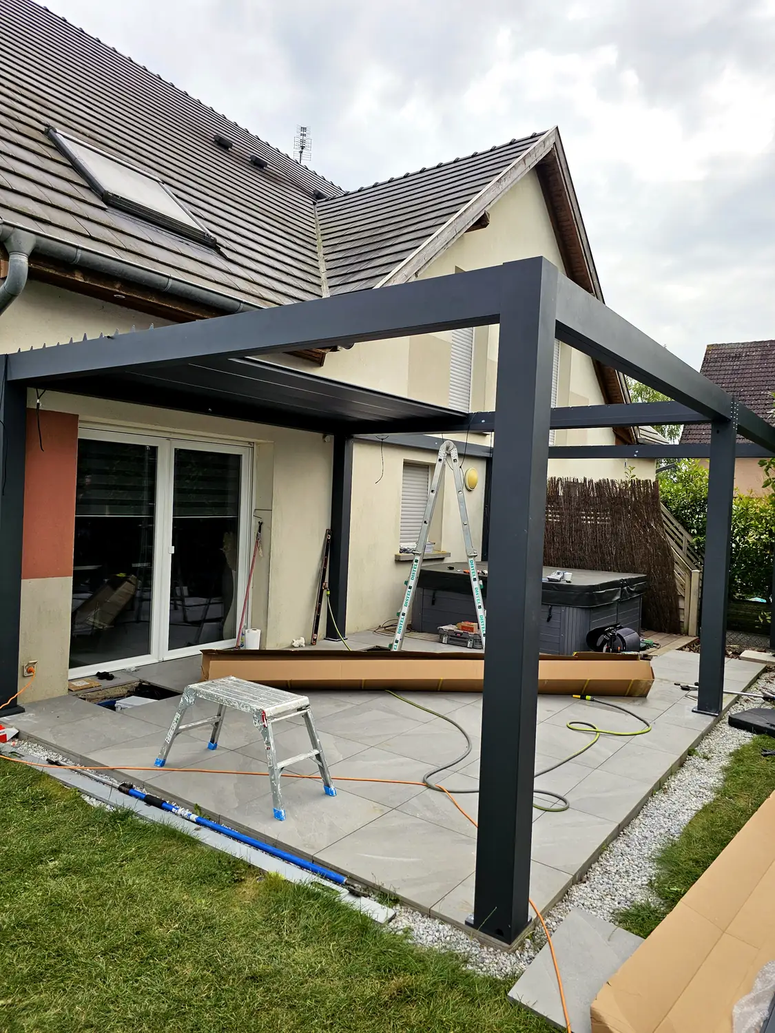 Pergola anthracite en cours de pose sur une terrasse dallée, échelle contre la façade, cartons et rallonges au sol, spa carré sombre à droite.